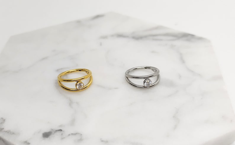 May include: Two simple rings with a single clear stone set in the center. The ring on the left is gold and the ring on the right is silver.