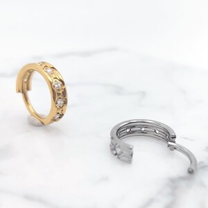 K&ouml;nnte beinhalten: Zwei Creolen mit einem dekorativen Design. Der Ohrring links ist golden mit klaren Steinen. Der Ohrring rechts ist silber mit klaren Steinen.