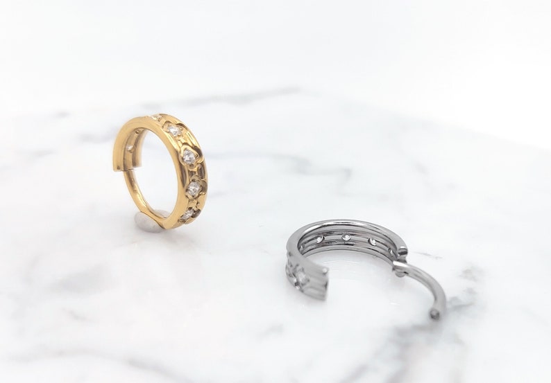 K&ouml;nnte beinhalten: Zwei Creolen mit einem dekorativen Design. Der Ohrring links ist golden mit klaren Steinen. Der Ohrring rechts ist silber mit klaren Steinen.