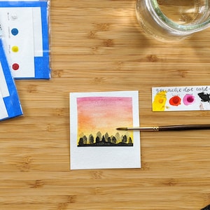 May include: A watercolor painting of a sunset over a forest. The painting is on a white card with a white border. The card is on a wooden surface. There is a paint palette with the words "gouache dot card" and the colors "golden yellow", "sapphire", "amid" and "onyx".