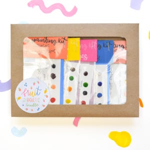 May include: A brown cardboard box with a clear plastic window showing a set of six gouache paint sets. Each set has a different color theme and includes a small card with the color name and a sample of the paint. The box has a sticker on the front that says "fruit basket bundle".