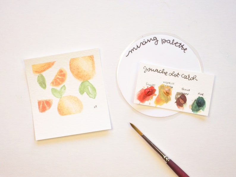 May include: A watercolor painting of oranges and leaves on a white card. A mixing palette with a gouache dot card showing four colors: scarlet, metallic cream, burnt umber, and pine.