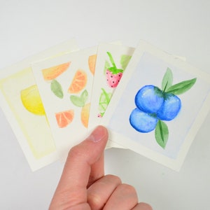 May include: Four watercolor paintings on white cardstock. The paintings depict a lemon, orange slices, strawberries, and blueberries.
