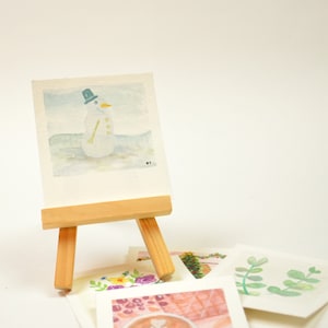 May include: A watercolour painting of a snowman with a blue hat and a yellow carrot nose, displayed on a small wooden easel. The painting is surrounded by other watercolour paintings on a white surface.