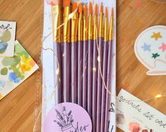 May include: A set of 10 purple paint brushes with orange bristles, packaged in a clear plastic bag. The packaging features the text "LAVENDER CHAI CO. brushes" and a logo with the text "Lavender Chai Co."