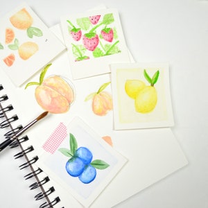 May include: A collection of watercolor paintings on white paper featuring various fruits, including lemons, peaches, blueberries, oranges, and strawberries. The paintings are arranged on a white surface with a spiral bound sketchbook and a paintbrush.