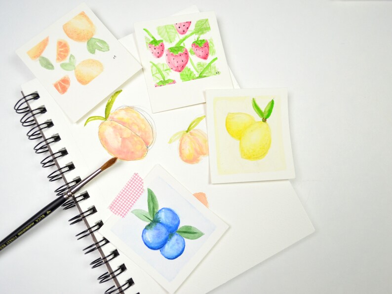 May include: A collection of watercolor paintings on white paper featuring various fruits, including lemons, peaches, blueberries, oranges, and strawberries. The paintings are arranged on a white surface with a spiral bound sketchbook and a paintbrush.