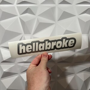 May include: A white sticker with the word "hellabroke" in black, outlined in a darker gray. The sticker is held in a hand against a white, geometric wall.