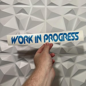May include: A rectangular adhesive decal featuring the text WORK IN PROGRESS in bold, blue, capital letters. The sticker is presented on a white backing paper, showcasing a clean, sans-serif font with sharp, angular edges for a modern appearance.