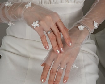 Peut inclure: Une paire de gants en tulle blanc avec de délicates fleurs blanches en guise d'embellissements. Les gants sont portés sur les mains d'une personne, avec une bague à un doigt.