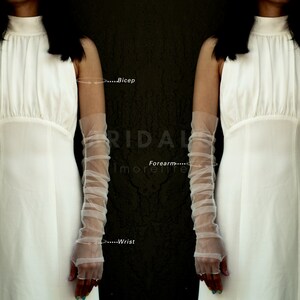 May include: A pair of white tulle elbow-length gloves with a gathered detail. The gloves are shown on a model wearing a white dress. The text "...Bicep", "...Forearm...", and "...Wrist" is visible on the image.