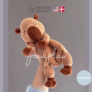 May include: A handmade, plush giraffe in shades of tan and brown. The crocheted giraffe has a rounded head, small ears, and stubby legs. The image includes the text "PATTERN OPSKRIFT" and "LOW-SEW".