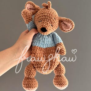 May include: A handmade crochet kangaroo plush toy in shades of brown and blue. The kangaroo has closed eyes, a black nose, and a light blue top. The ears and inner ears are a lighter shade of brown. The toy is held up by a hand.