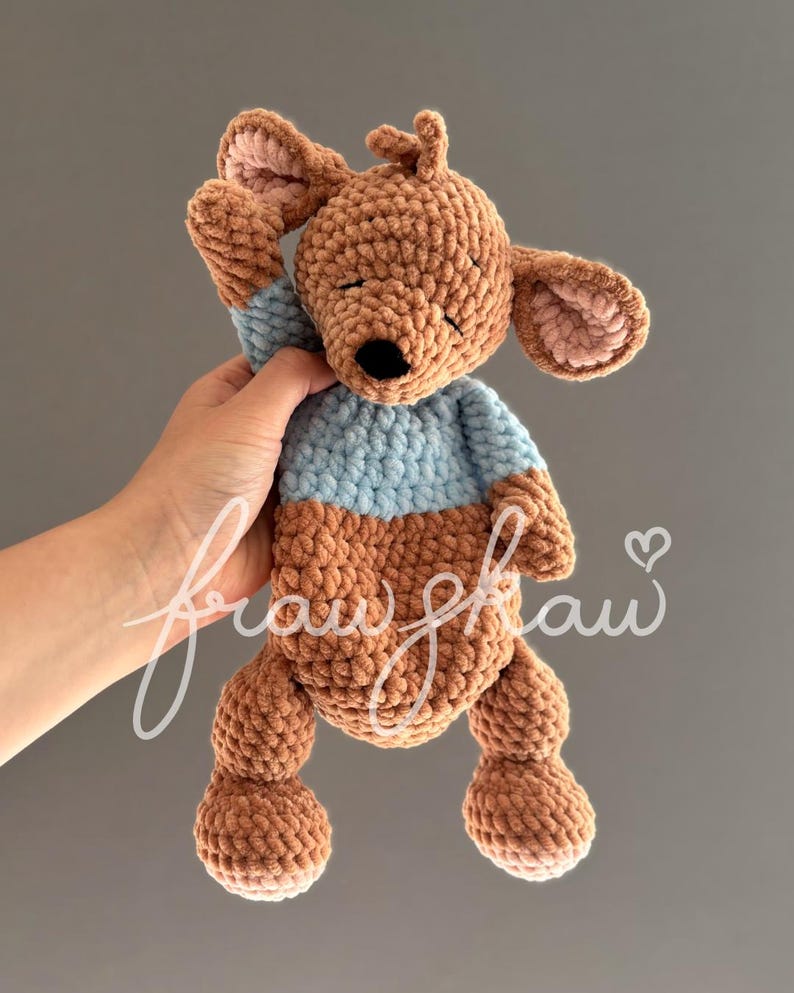 May include: A handmade crochet kangaroo plush toy in shades of brown and blue. The kangaroo has closed eyes, a black nose, and a light blue top. The ears and inner ears are a lighter shade of brown. The toy is held up by a hand.