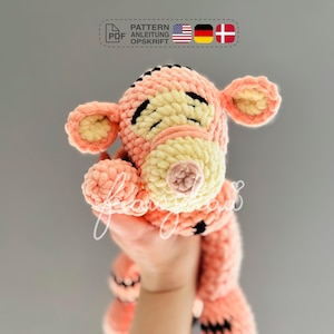 Op de afbeelding: Een gehaakt roze en crèmekleurig Tigger knuffel met zwarte ogen en een zwarte neus. Het speelgoed wordt vastgehouden door een hand.
