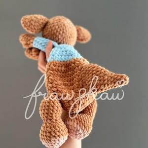 May include: A handmade brown crochet doll with a light blue scarf. The doll has large ears and is held up by a hand. The doll's feet are a lighter shade of brown. The background is a solid gray.