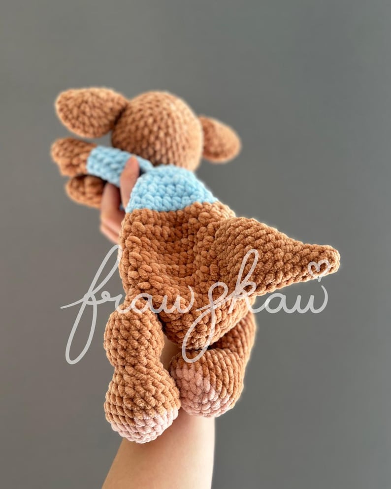 May include: A handmade brown crochet doll with a light blue scarf. The doll has large ears and is held up by a hand. The doll's feet are a lighter shade of brown. The background is a solid gray.