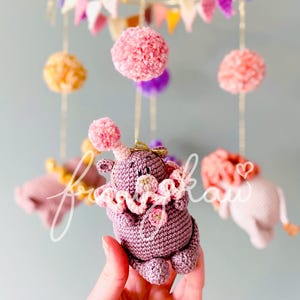 Puede incluir: M&oacute;vil de beb&eacute; hecho a mano con ganchillo, con un hipop&oacute;tamo morado, cuello con volantes y pompones rosas. El m&oacute;vil incluye pompones rosas, naranjas y morados, y peque&ntilde;os elefantes de ganchillo. Colgado de hilos dorados.