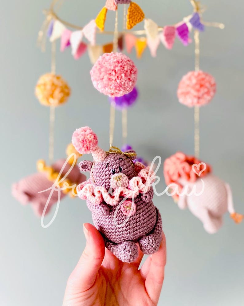 Puede incluir: M&oacute;vil de beb&eacute; hecho a mano con ganchillo, con un hipop&oacute;tamo morado, cuello con volantes y pompones rosas. El m&oacute;vil incluye pompones rosas, naranjas y morados, y peque&ntilde;os elefantes de ganchillo. Colgado de hilos dorados.
