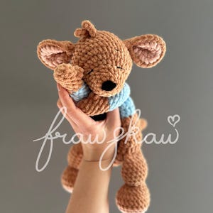 May include: A handmade brown crochet kangaroo plush toy with a light blue shirt. The kangaroo has large ears with pink inner ears and a black nose. The plush is held up against a gray background.