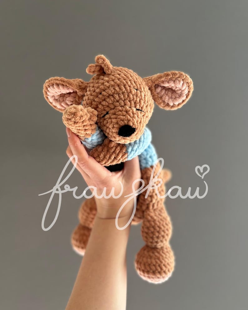 May include: A handmade brown crochet kangaroo plush toy with a light blue shirt. The kangaroo has large ears with pink inner ears and a black nose. The plush is held up against a gray background.
