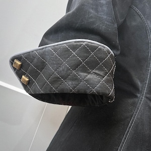 May include: Close-up of a black coat sleeve. The cuff features a quilted pattern with white stitching and is trimmed with a gray border. Two gold-colored square buttons adorn the cuff. The coat's fabric has a slightly distressed appearance.