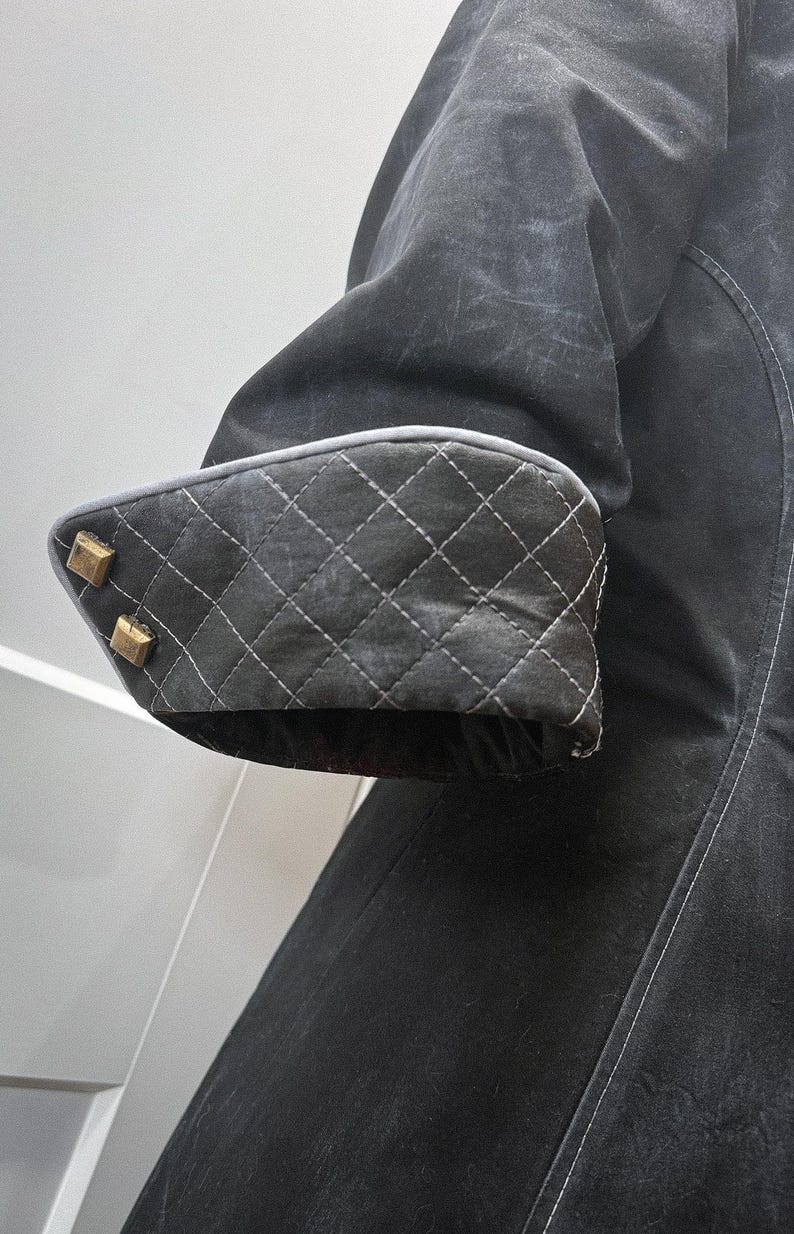 May include: Close-up of a black coat sleeve. The cuff features a quilted pattern with white stitching and is trimmed with a gray border. Two gold-colored square buttons adorn the cuff. The coat's fabric has a slightly distressed appearance.