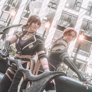 May include: Two people in cosplay attire, holding large, ornate weapons. They wear black and gold outfits with intricate details, including armor-like elements and decorative patterns. The background features a modern cityscape with buildings and streetlights.