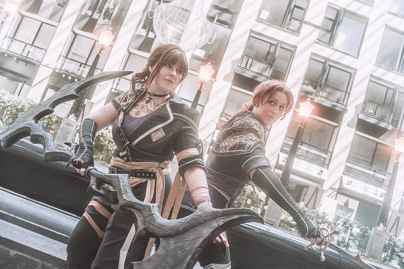 May include: Two people in cosplay attire, holding large, ornate weapons. They wear black and gold outfits with intricate details, including armor-like elements and decorative patterns. The background features a modern cityscape with buildings and streetlights.