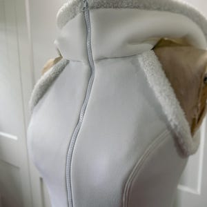 May include: A white hooded vest with a full-length zipper. The hood and collar are lined with a soft, white, fuzzy material. The vest is displayed on a mannequin.