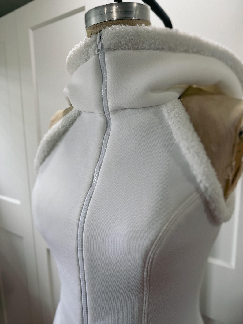 May include: A white hooded vest with a full-length zipper. The hood and collar are lined with a soft, white, fuzzy material. The vest is displayed on a mannequin.