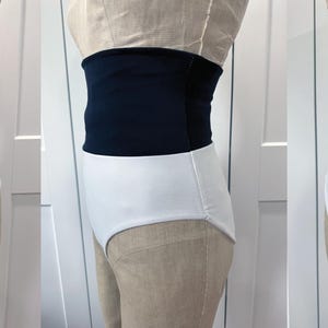 May include: A two-piece swimsuit with a high-waisted design. The top is a solid navy blue, and the bottoms are white. The swimsuit is displayed on a mannequin, showcasing the front, side, and back views of the garment.