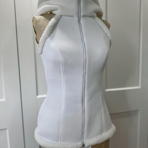 May include: A white sleeveless vest with a hood and a full-length zipper. The vest has a fitted silhouette with faux fur trim around the hood, armholes, and hemline. The garment is displayed on a mannequin.