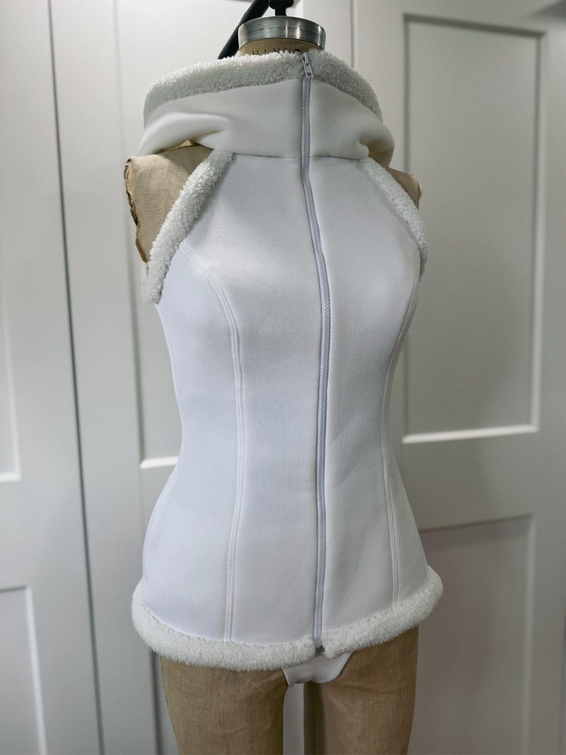 May include: A white sleeveless vest with a hood and a full-length zipper. The vest has a fitted silhouette with faux fur trim around the hood, armholes, and hemline. The garment is displayed on a mannequin.