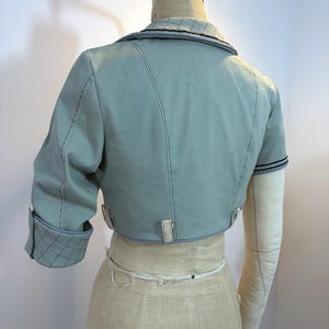 May include: A light blue cropped jacket with short sleeves, displayed on a dress form. The jacket features contrasting stitching, a notched collar, and a quilted pattern on the sleeve cuffs. The dress form is beige.
