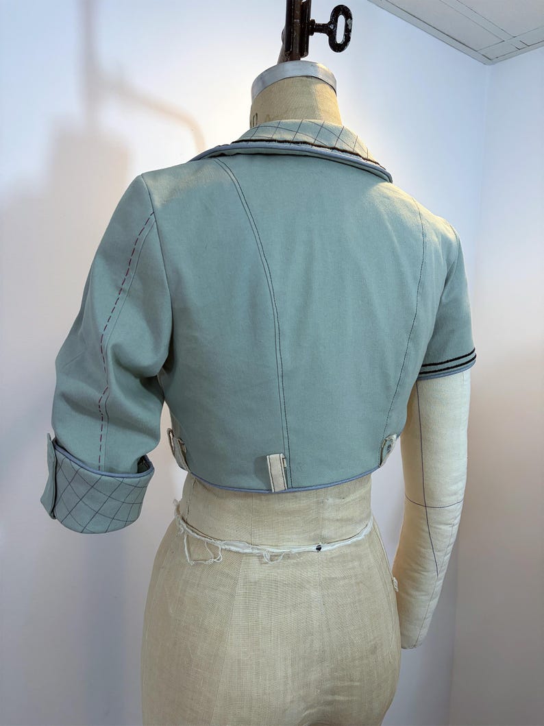 May include: A light blue cropped jacket with short sleeves, displayed on a dress form. The jacket features contrasting stitching, a notched collar, and a quilted pattern on the sleeve cuffs. The dress form is beige.