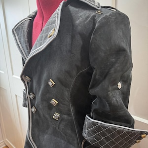 May include: A dark gray coat with a double-breasted design, featuring gold-colored square buttons and a quilted collar and cuffs. The coat has a tailored fit and a contrasting white trim along the edges. The coat is displayed on a red dress form.