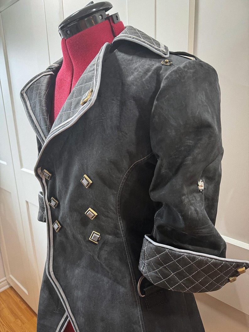 May include: A dark gray coat with a double-breasted design, featuring gold-colored square buttons and a quilted collar and cuffs. The coat has a tailored fit and a contrasting white trim along the edges. The coat is displayed on a red dress form.