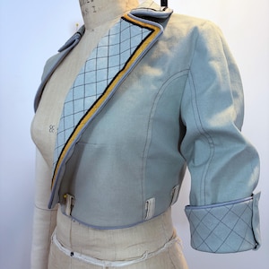 May include: A light blue cropped jacket with a quilted collar and cuffs. The collar features a diamond pattern with yellow and black trim. The jacket has three-quarter sleeves and is displayed on a dress form.