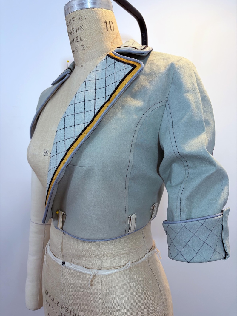 May include: A light blue cropped jacket with a quilted collar and cuffs. The collar features a diamond pattern with yellow and black trim. The jacket has three-quarter sleeves and is displayed on a dress form.