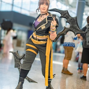 May include: A person in a cosplay outfit, featuring a black and gold jacket, gray pants, and black boots. The outfit includes a yellow sash and wristbands. The person is holding a dark, stylized weapon.