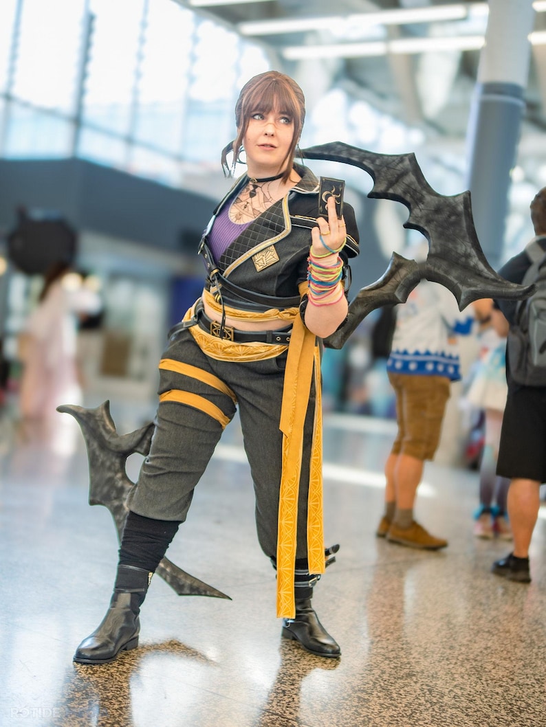 May include: A person in a cosplay outfit, featuring a black and gold jacket, gray pants, and black boots. The outfit includes a yellow sash and wristbands. The person is holding a dark, stylized weapon.