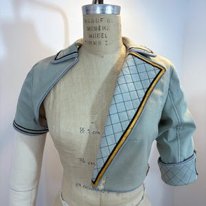 May include: A light blue cropped jacket with a quilted pattern on the lapel and cuffs. The jacket has black and gold trim and is displayed on a mannequin. The mannequin has measurements of 88.5 cm, 76 cm, and 67 cm.