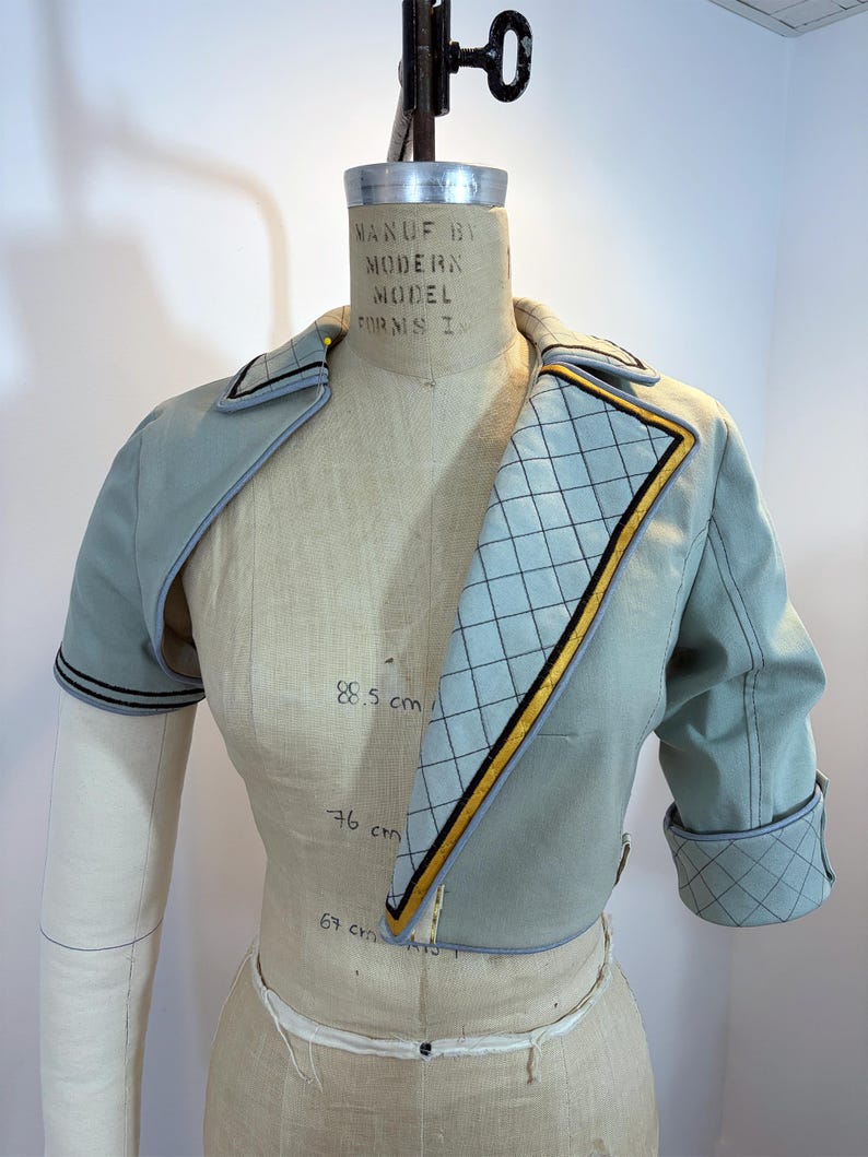 May include: A light blue cropped jacket with a quilted pattern on the lapel and cuffs. The jacket has black and gold trim and is displayed on a mannequin. The mannequin has measurements of 88.5 cm, 76 cm, and 67 cm.