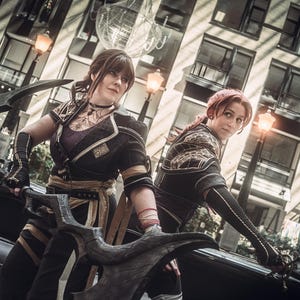 May include: Two individuals in cosplay attire, posing with large, ornate weapons. They wear black and gold outfits with intricate detailing, set against a backdrop of a modern building with street lamps. The overall aesthetic suggests a fantasy or action theme.