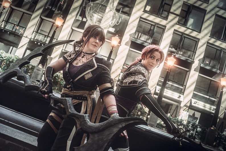 May include: Two individuals in cosplay attire, posing with large, ornate weapons. They wear black and gold outfits with intricate detailing, set against a backdrop of a modern building with street lamps. The overall aesthetic suggests a fantasy or action theme.