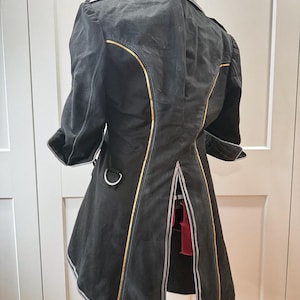 May include: Black coat with gold trim and silver accents. The coat has a unique design with a split back and three-quarter sleeves. The coat is hanging on a hanger against a white background.