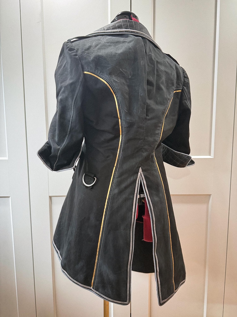 May include: Black coat with gold trim and silver accents. The coat has a unique design with a split back and three-quarter sleeves. The coat is hanging on a hanger against a white background.