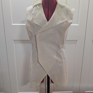May include: A sleeveless, cream-colored vest with a shawl collar and an asymmetrical front closure. The vest is displayed on a red dress form and features visible stitching along the edges. The garment appears to be made of a light, textured fabric.