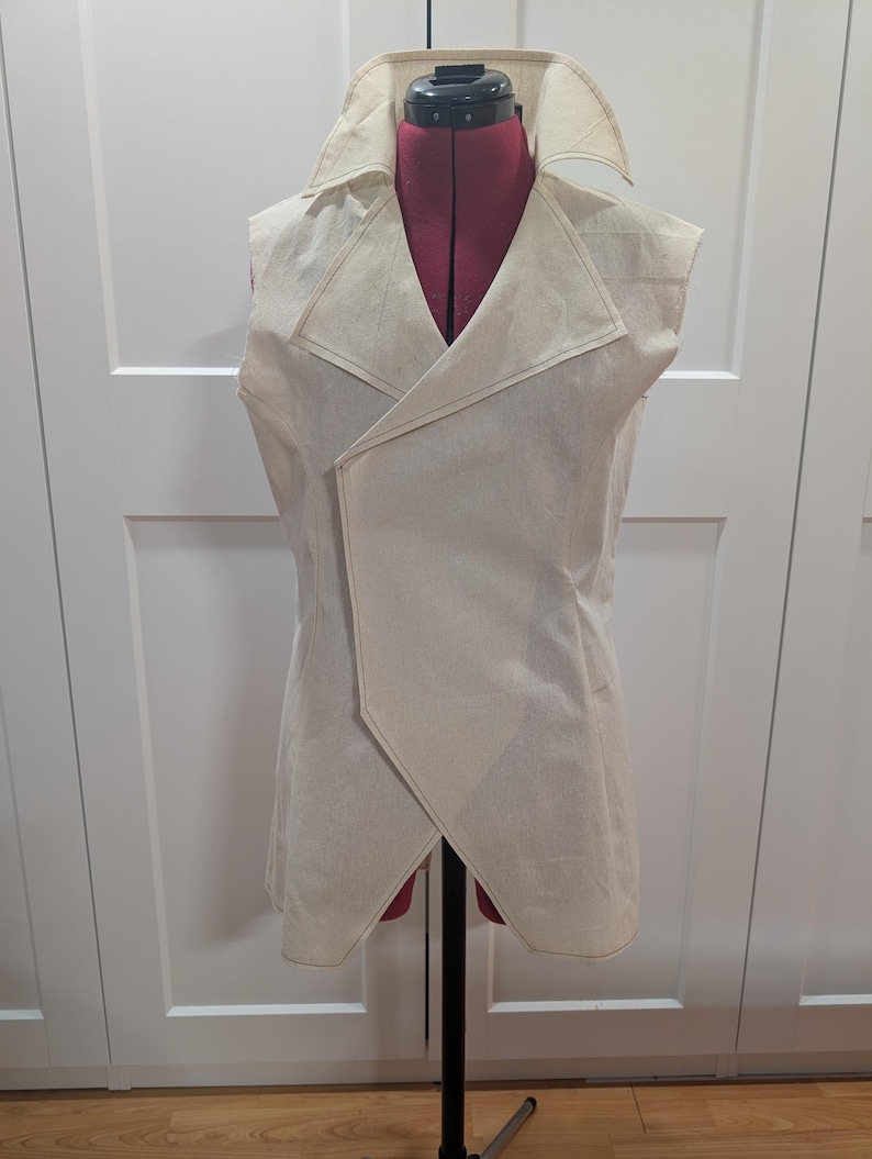 May include: A sleeveless, cream-colored vest with a shawl collar and an asymmetrical front closure. The vest is displayed on a red dress form and features visible stitching along the edges. The garment appears to be made of a light, textured fabric.
